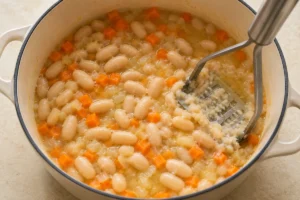 A potato masher thickening simmered white beans and carrot in vegetable broth